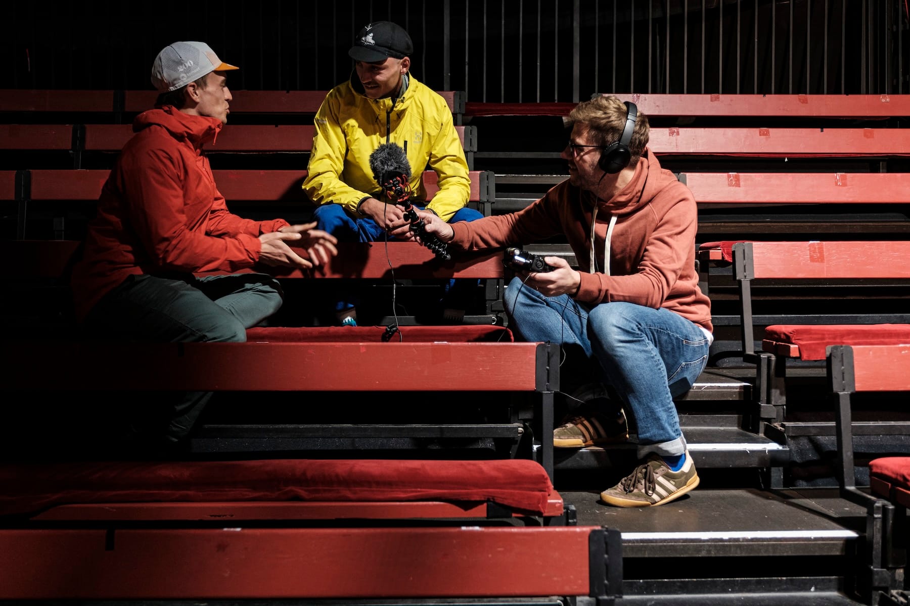 Iain Broome with headphones on holding out a microphone to interview a cyclist. They are sitting on rows of empty red bleacher-style seating. 