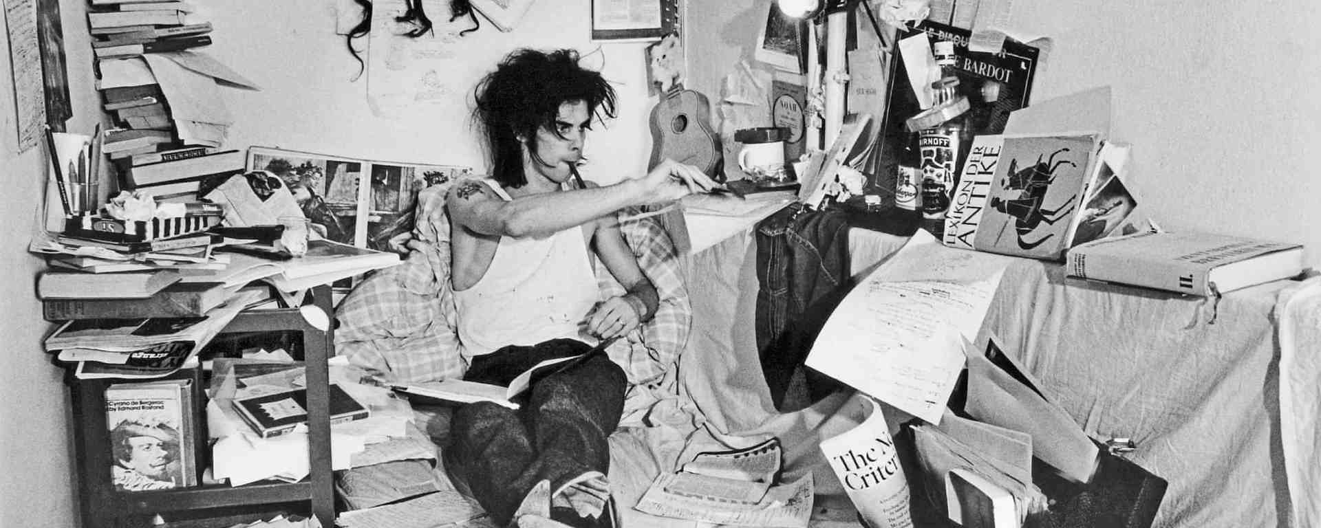 Nick Cave writing in a cluttered area full of books, papers and cushions