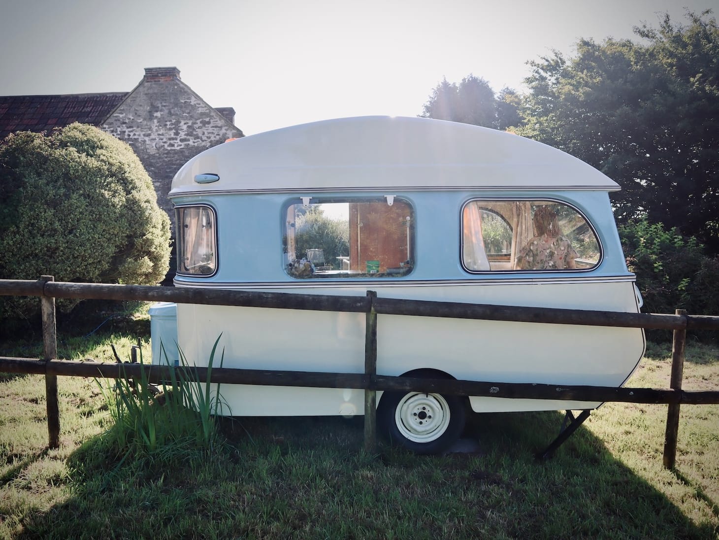 Small white caravan parked on grass with a pale blue stripe across the middle. A stone cottage and trees in the background. 